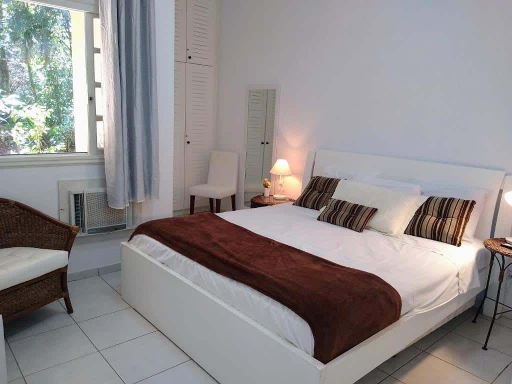 Quarto em Riviera Flat Service- 109, uma cama de casal, um armário, uma cadeira, uma mesinha de cabeceira com abajur, um ar-condicionado, uma poltrona e uma janela com cortinas, para representar pousadas em Riviera de São Lourenço.