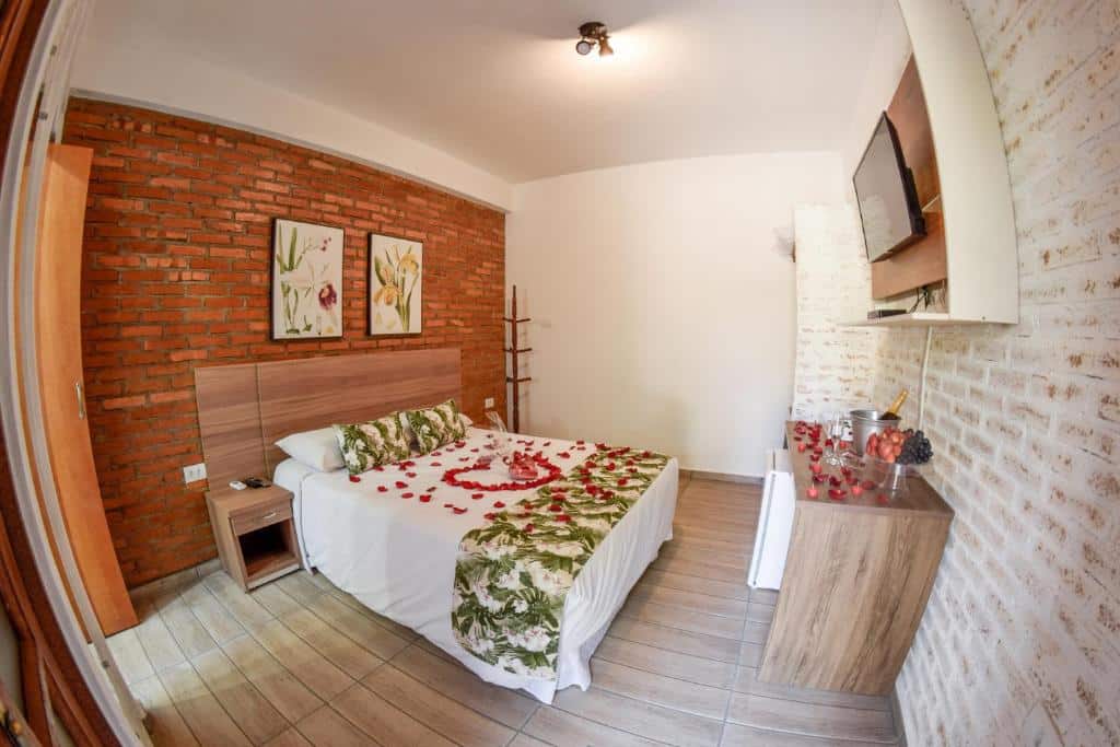 Quarto da pousada do Vale das Orquídeas, para ilustrar o post sobre pousadas perto de Ribeirão Preto. No quarto vemos uma cama box de casal decorada com pétalas de rosas, e mesa em sua frente também está decorada da mesma forma, possuindo um balde de champagne e frutas em uma travessa. Uma televisão está de frente para a cama, fixa em um suporte na parede. Debaixo da mesa, há um frigobar. Dois quadros decoram a mesma parede que a cama está encostada.