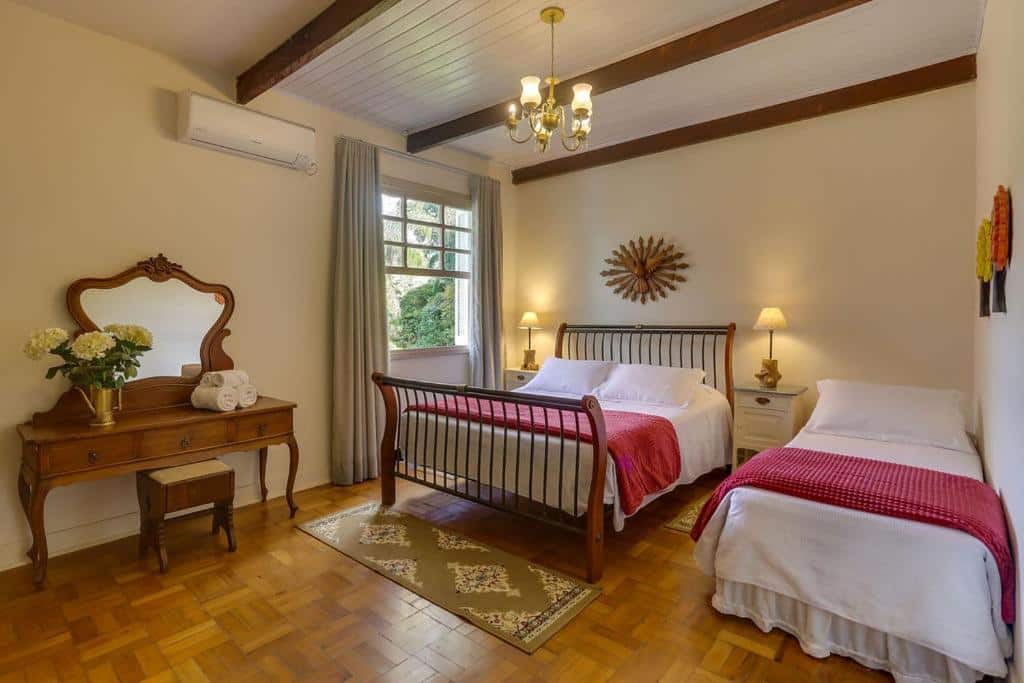 Imagem do quarto da Pousada Famiglia Bartho, em Espírito Santo do Pinhal, para representar o post sobre pousadas perto de Ribeirão Preto. A cama é espaçosa e de casal, nos dois lados dela há uma mesa de cabeceira com abajur em cima. Também há outra cama de solteiro no quarto, do lado direito da cama de casal. Do lado esquerdo, está a janela, e uma penteadeira de madeira. 