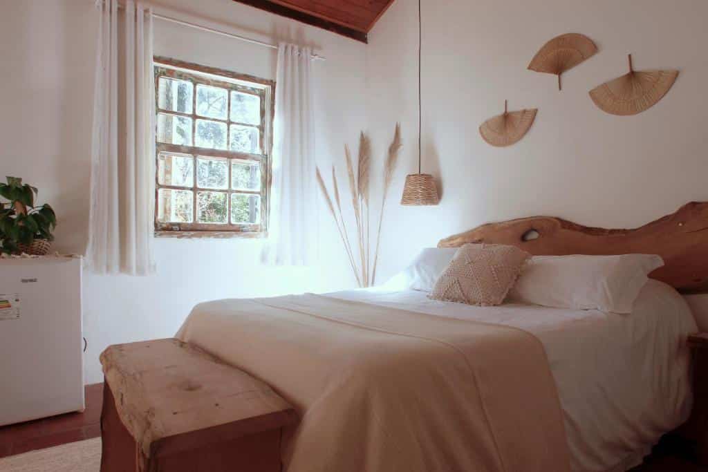 Quarto da Hospedagem Fazenda Nova América, em Brotas, para representar o post sobre pousadas perto de Ribeirão Preto. Os detalhes da suíte são de madeira clara. A cama é de casal, há uma janela do seu lado esquerdo e um banco em sua frente. Um frigobar também está disponível.