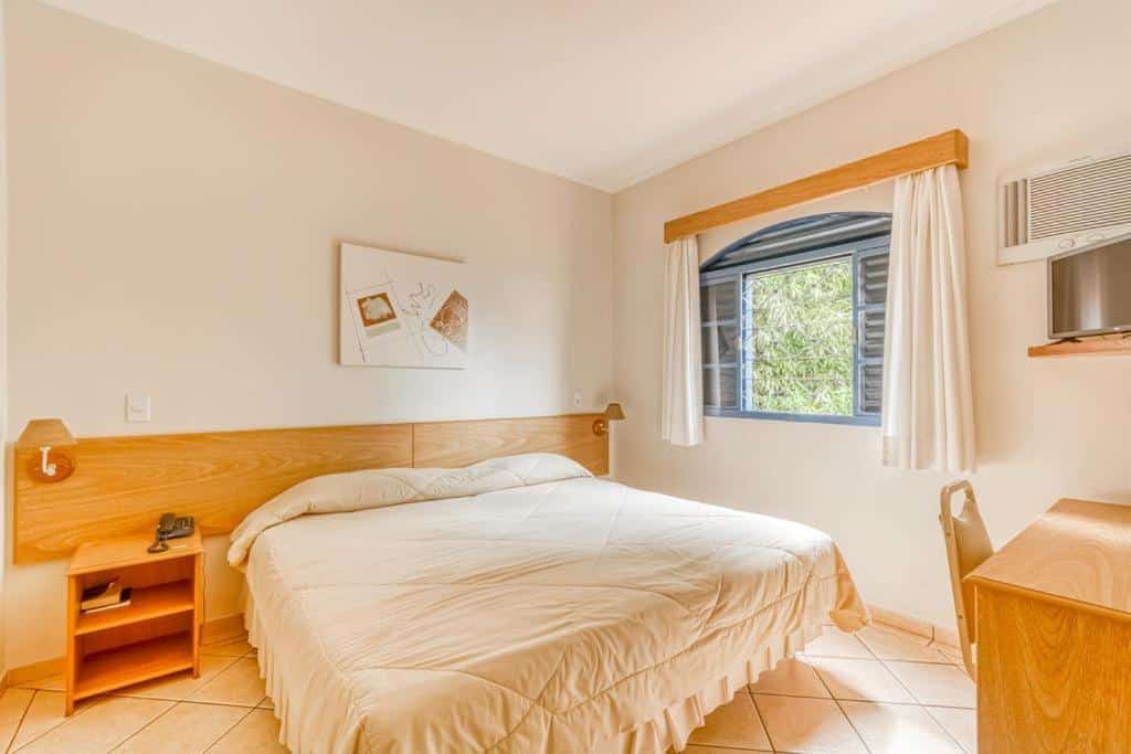 Foto do quarto do Jardin Park Hotel, em Jardinópolis, para representar o post sobre pousadas perto de Ribeirão Preto. A suíte possui detalhes de madeira clara, a cama é de casal e possui uma mesa de cabeceira do seu lado esquerdo. Do lado direito, há uma janela. Vemos uma escrivaninha de madeira na frente da cama, e uma TV pousada sobre uma prateleira.