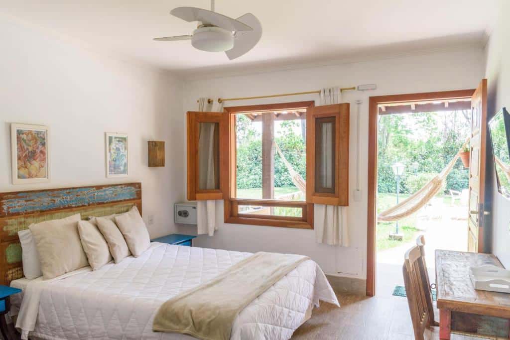 Imagem do Hotel Recanto da Cachoeira, em Socorro, para ilustrar o post sobre pousadas perto de Ribeirão Preto. O quarto possui decoração rústica, a acama é de casal e possui mesas de cabeceira em ambos os lados. Do seu lado direito, há uma janela e porta que leva para a área externa da pousada. De frente para a cama, há uma escrivaninha e um televisão na parede.