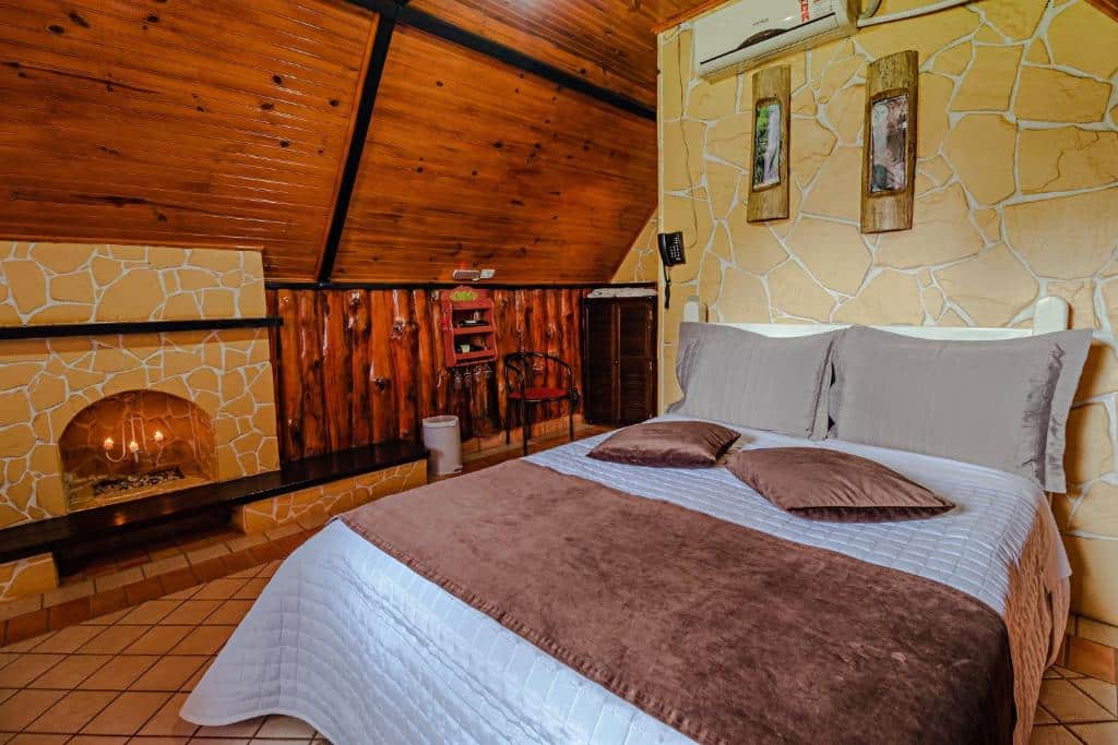 Foto do quarto do Mirágua Refugios Pousada, em Brotas, para representar o post sobre pousadas perto de Ribeirão Preto. A cama é de casal e possui almofadas e travesseiros. Do lado esquerdo, há uma lareira e decorações de madeira. É possível ver um ar condicionado na parte superior da foto, na mesma parede em que a cama está encostada.