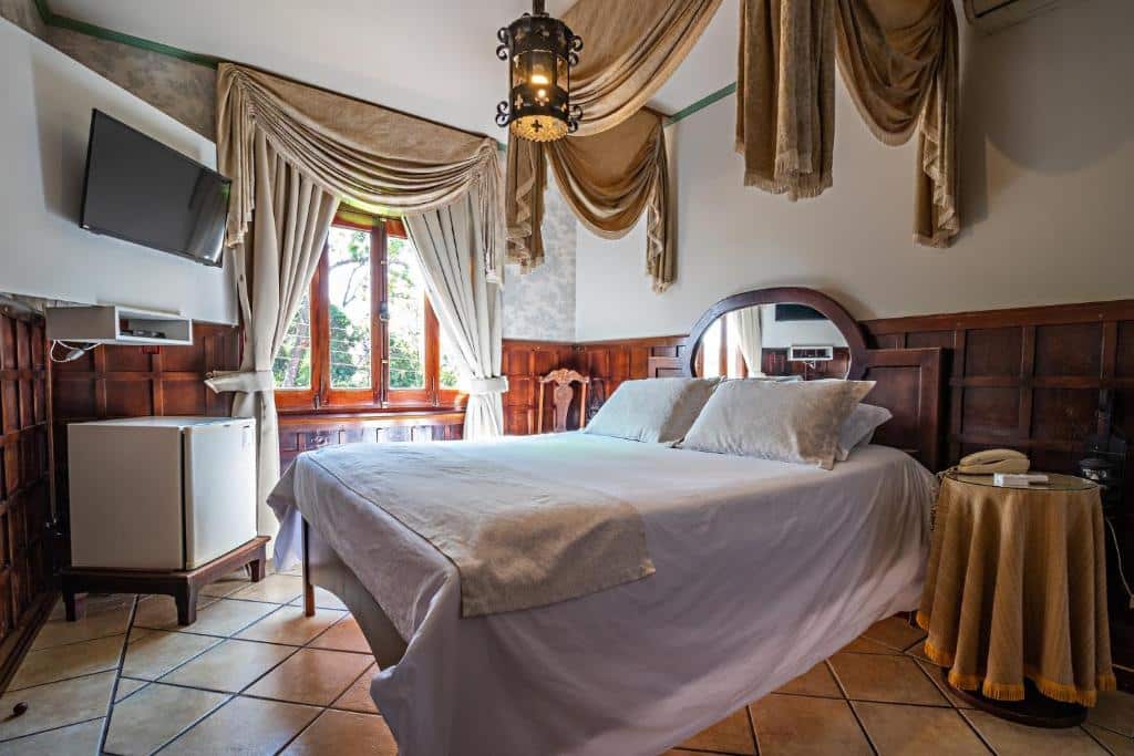 Imagem de um dos quartos da Pousada Estalagem de Brotas, em Brotas, para ilustrar o post sobre pousadas perto de RIbeirão Preto. O quarto possui uma cama de casal, as paredes são decoradas até a metade em painéis de madeira e muitas cortinas enfeitam o ambiente. Possui uma janela e de seu lado há uma TV voltada para a cama, e embaixo tem um frigobar.