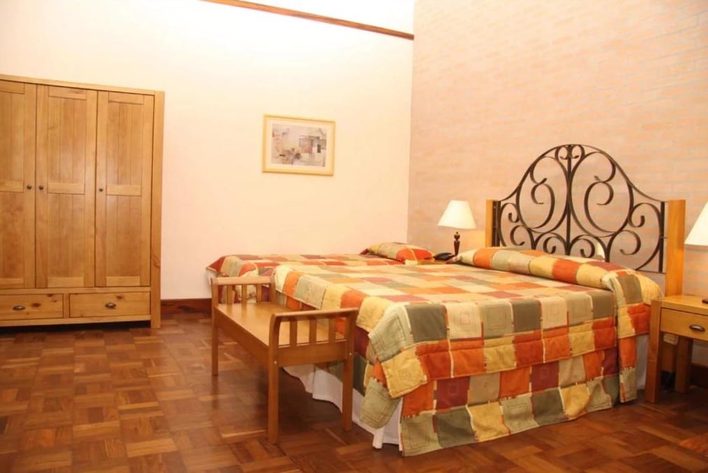 Foto do Hotel Fazenda Salto Grande, em Araraquara, para representar o post sobre pousadas em Ribeirão Preto. A cama é de casal, e possui uma mesa de cabeceira com abajur no lardo esquerdo e direito. Ao lado da cama vemos também um guarda-roupa de madeira e um quadro ao lado na parede.