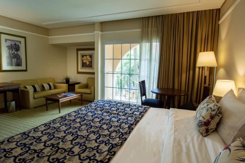 Imagem do Royal Palm Plaza Resort, em Campinas, para ilustrar o post sobre pousadas perto de Ribeirão Preto. A cama é de casal, do seu lado há uma mesa de cabeceira com abajur. A janela do quarto dá acesso para uma varanda. Há um sofá e uma poltrona com uma mesa de centro de madeira n frente. Quadros decoram a suíte.