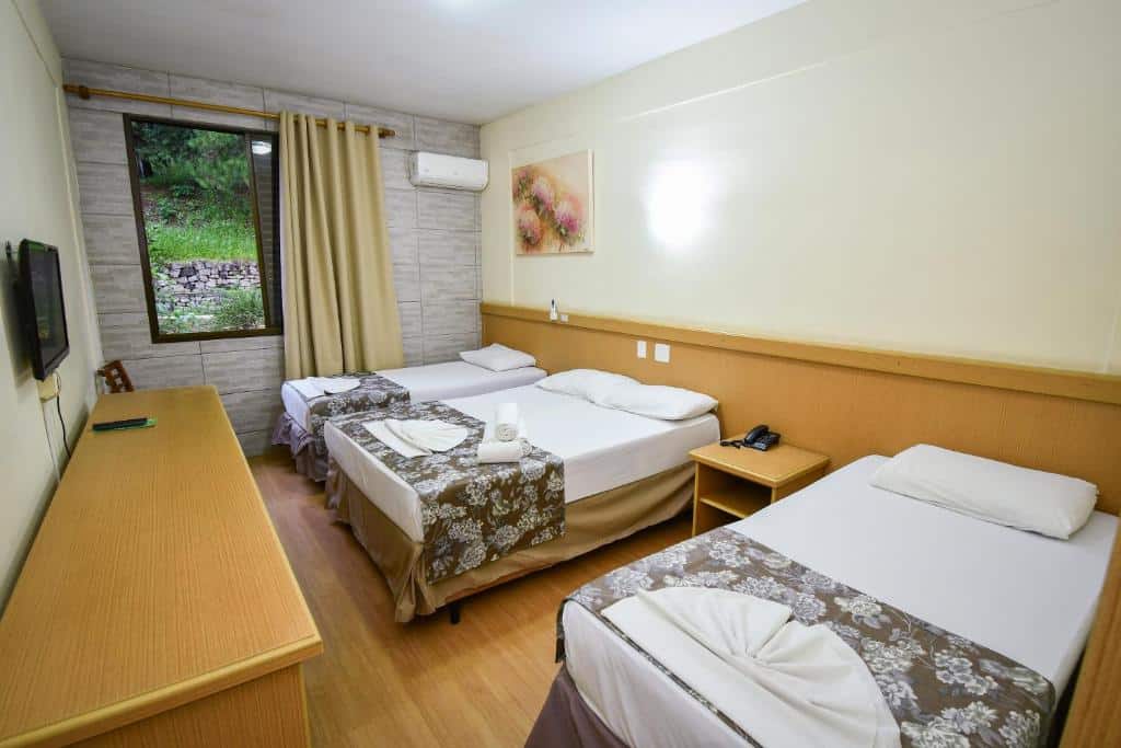 Foto do Vilage Inn All Inclusive Poços de Caldas, para representar o post sobre pousadas perto de Ribeirão Preto. Há uma cama box de casal entre outras duas camas de solteiro. Na frente, há uma mesa de televisão, e a TV está fixa na parede. O quarto possui uma janela e um ar-condicionado do lado. 