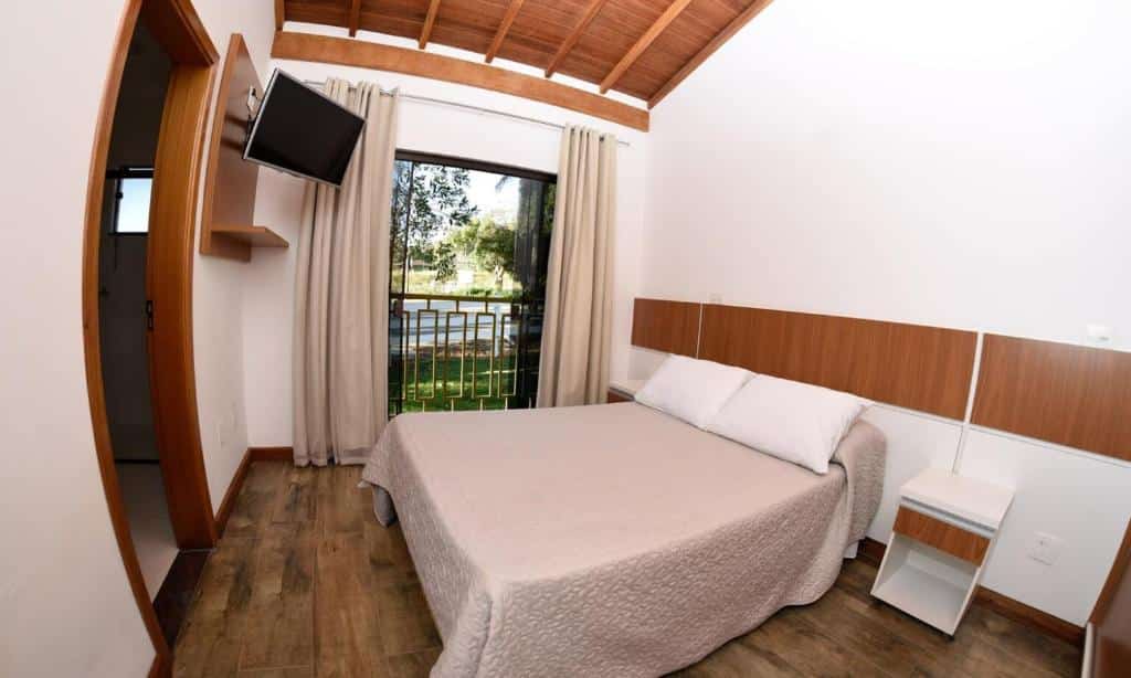 Foto do Hotel Fazenda, em Poços de Caldas, para representar o post sobre pousadas perto de Ribeirão Preto. O quarto possui detalhes de madeira, a cama é box e de casal, do seu lado esquerdo há uma porta que possui vista para a piscina. Há um banheiro privativo e uma televisão fixa na parede, voltada para a cama.