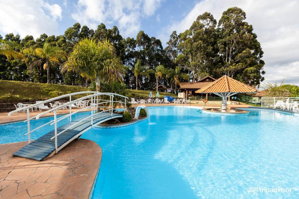 Foto da piscina do Hotel Fazenda, em Poços de Caldas. Ela é grande e ocupa um espaço enorme do resort, possui uma ponte sobre a água para um pequeno jardim. Um quiosque está posto no meio da piscina. Em volta, há muitas espreguiçadeiras, e também podemos ver diversas árvores no fundo.