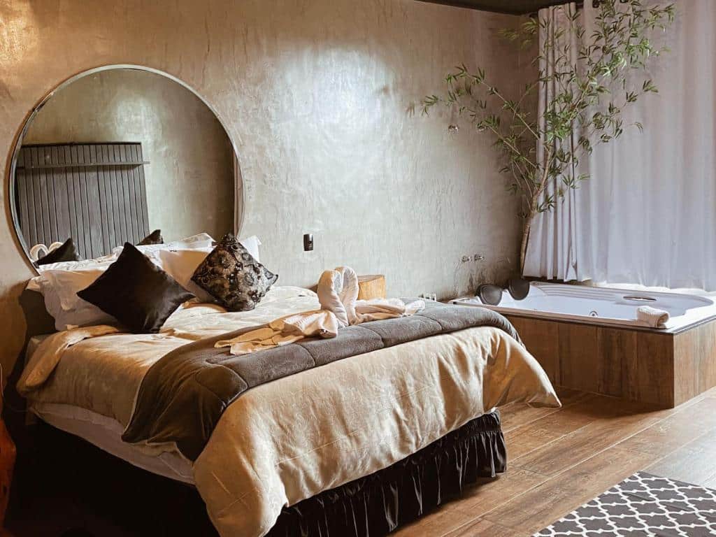 Quarto da Pousada Café Poesia. No lado esquerdo é possível ver uma cama de casal, com um grande espelho circular na parede de trás. Do lado direito do quarto há uma banheira, com uma planta que enfeita a área. Imagem utilizada para ilustrar o post pousadas pet friendly em Campos do Jordão.