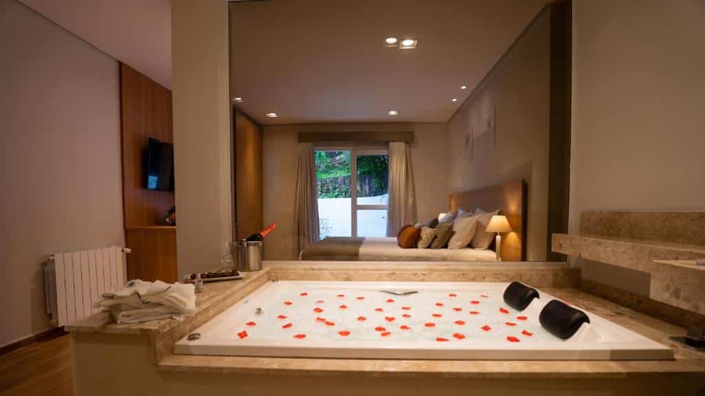 Quarto da Pousada Grandchamp. Há uma banheira com pétalas de rosas em primeiro plano. Ao fundo é possível ver a cama do lado direito, com abajures dos dois lados e uma televisão do lado esquerdo. Ainda mais ao fundo está uma porta com cortinas, que dá acesso a parte externa do quarto. Imagem utilizada para ilustrar o post pousadas pet friendly em Campos do Jordão.