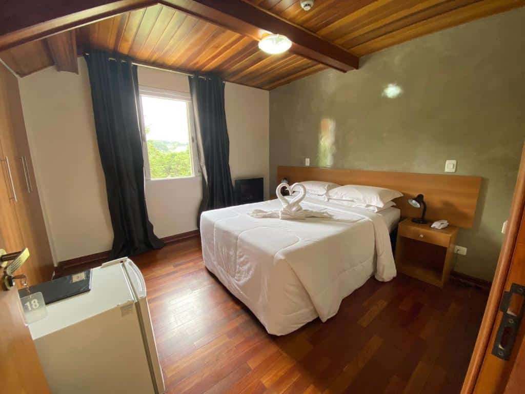 Quarto da Pousada Jordão. Há uma cama de casal centralizada com mesas de cabeceira e abajures dos dois lados e em frente a cama é possível ver um frigobar. No fundo está uma janela com cortinas. Imagem utilizada para ilustrar o post pousadas pet friendly em Campos do Jordão.
