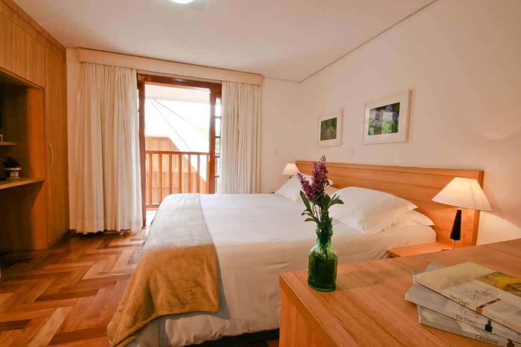 Quarto da Pousada Campos dos Holandeses. Do lado direito há uma cama de casal, com abajures dos dois lados. Do lado esquerdo pode-se ver armários e ao fundo uma porta com cortinas, que dá acesso a varanda. Imagem utilizada para ilustrar o post pousadas pet friendly em Campos do Jordão.