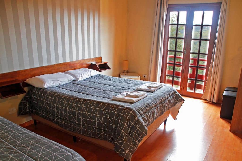 Quarto da Pousada AlpenRose. É possível ver uma cama de casal centralizada e um frigobar e um abajur no lado esquerdo. Ao fundo há uma porta com cortinas, que dá acesso a varanda do quarto. Imagem utilizada para ilustrar o post pousadas pet friendly em Campos do Jordão.