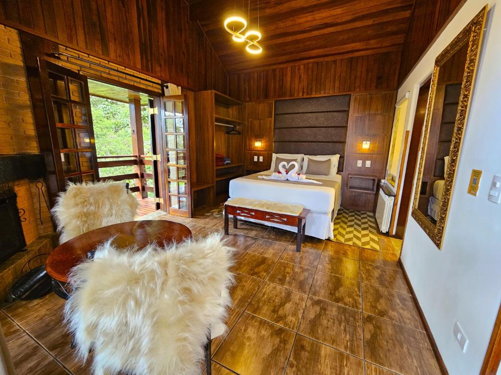 Quarto da Pousada Recanto dos Sonhos. No centro do quarto há uma cama de casal, com abajures dos dois lados e um armário ao lado direito. Na frente da cama há um pequeno banco e ainda mais a frente uma mesa com duas grandes cadeiras felpudas. Do lado esquerdo do quarto há um espelho e uma porta e do lado direito duas portas que dão acesso a varanda. Imagem utilizada para ilustrar o post pousadas pet friendly em Campos do Jordão.