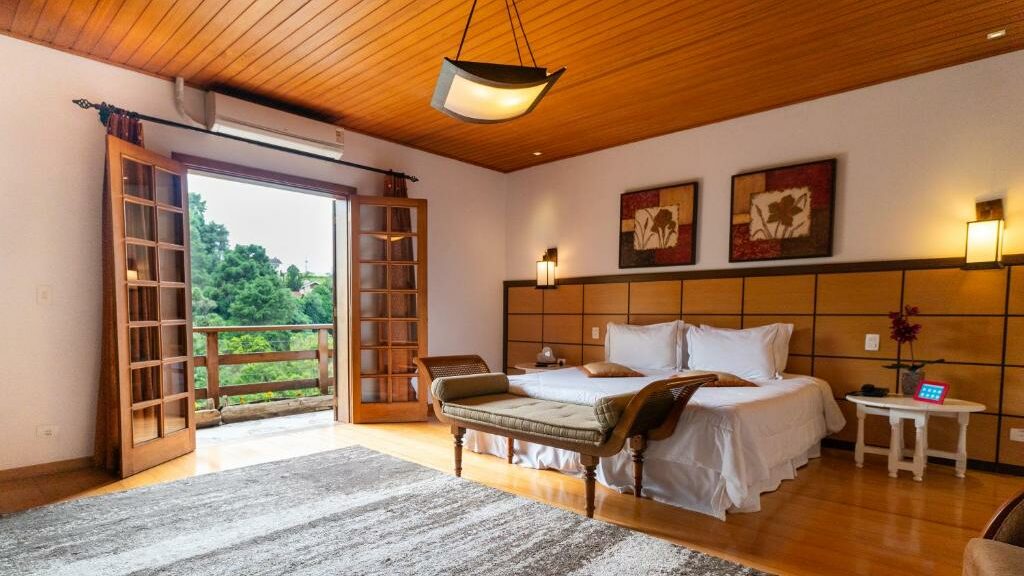 Quarto da Pousada Apple House. No lado direito da imagem há uma cama de casal, com mesas de cabeceira e abajures dos dois lados e quadros na parede de trás. Logo a frente da cama está um divã. Ao fundo é possível ver as portas que vão para a varanda do quarto. Imagem utilizada para ilustrar o post pousadas pet friendly em Campos do Jordão.