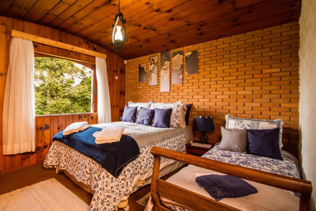 Quarto da Pousada Déjà vu. No lado direito há uma de solteiro e no lado esquerdo uma cama de casal. No espaço entre as camas há uma mesa de cabeceira com abajur. No fundo do quarto há uma janela com cortinas. Imagem utilizada para ilustrar o post pousadas pet friendly em Campos do Jordão.