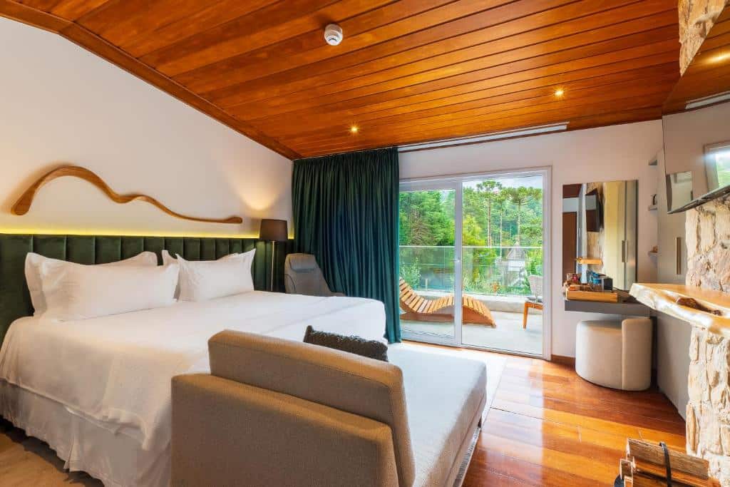 Quarto da Pousada Boutique Figueira da Serra. Há uma cama de casal centralizada, uma poltrona e um abajur no lado esquerdo da cama e em frente a cama há um pequeno sofá. Na parede ainda em frente a cama há uma televisão. Ao fundo pode-se ver um espelho com banquinho e uma porta com cortinas que vai para a varanda. Imagem utilizada para ilustrar o post pousadas pet friendly em Campos do Jordão.
