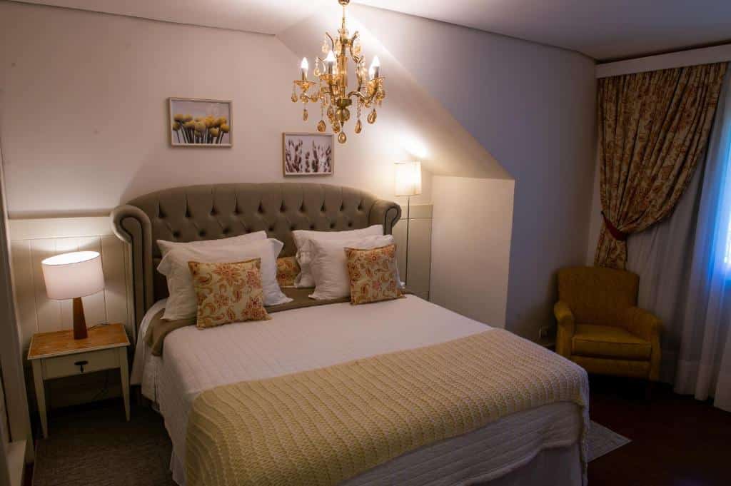 Quarto da Pousada das Lavandas. No centro da imagem há uma cama, com abajures dos dois lados e um lustre acima. No lado direito há uma poltrona e uma janela com cortinas. Imagem utilizada para ilustrar o post pousadas pet friendly em Campos do Jordão.