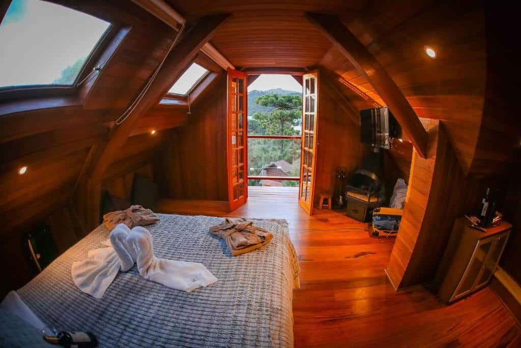 Quarto da Mansão Austríaca Guesthouse. Na esquerda é possível ver uma cama de casal e algumas janelas, a direita há uma televisão em cima de uma lareira e ao fundo observa-se uma porta que dá acesso a varanda do quarto. Imagem utilizada para ilustrar o post pousadas pet friendly em Campos do Jordão.