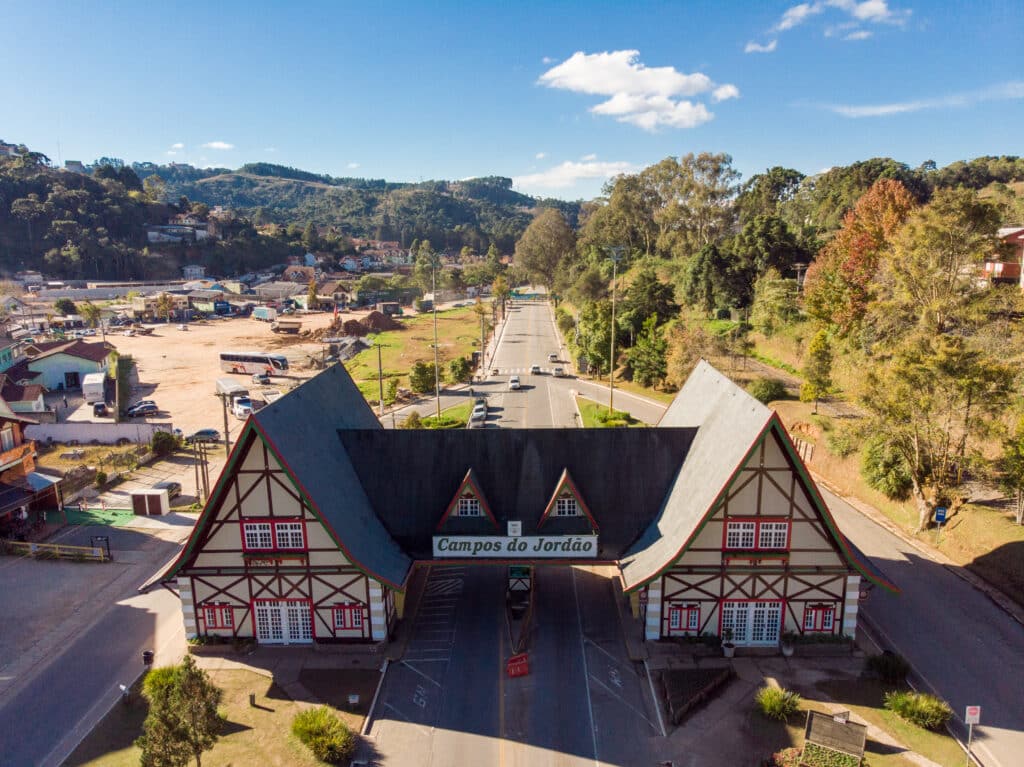 Entrada da cidade de Campos do Jordão vista de cima. No centro da imagem há uma estrutura com duas casas em estilo europeu ligadas por uma estrutura em seus tetos. Entre essa entrada se passa uma avenida de mão dupla e no lado direito há várias árvores.
