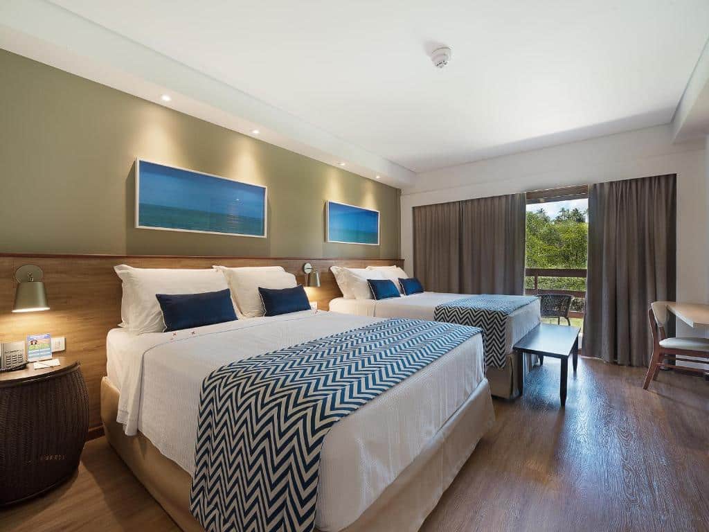 suíte do Salinas Maragogi All Inclusive Resort com duas camas de casal, uma mesa para trabalho e uma varanda com vista para a vegetção