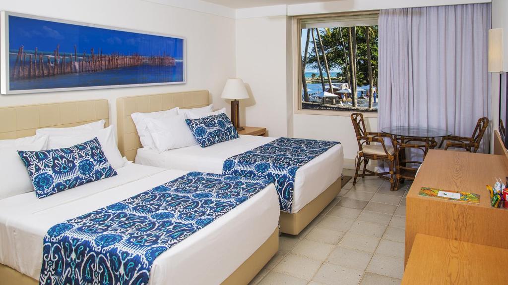 suíte do Jatiuca Hotel & Resort com duas camas de casal, uma mesa para dois uma janela com vista para a praia