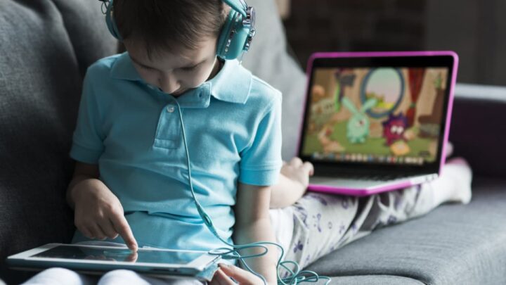 No primeiro plano, criança a usar um tablet, com auscultadores; no segundo plano, criança a usar um computador.