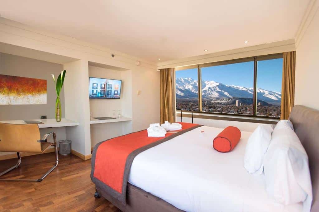 Quarto do Boulevard Suites Ferrat  com cama de casal do lado direito com mesa de trabalho a frente da cama e do lado esquerdo do quarto janelas amplas com vista para a cidade e montanhas.