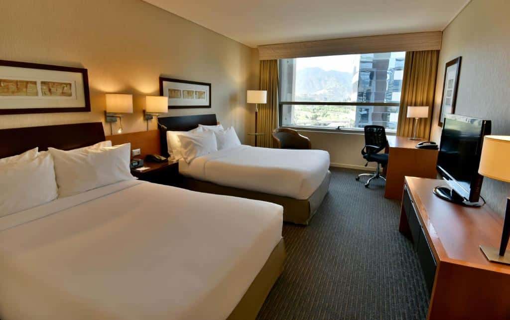 Quarto do Holiday Inn Express Santiago Las Condes, an IHG Hotel com duas camas de casal do lado esquerdo, em frente as camas uma cômoda com TV e do lado esquerdo do quarto uma mesa de trabalho.