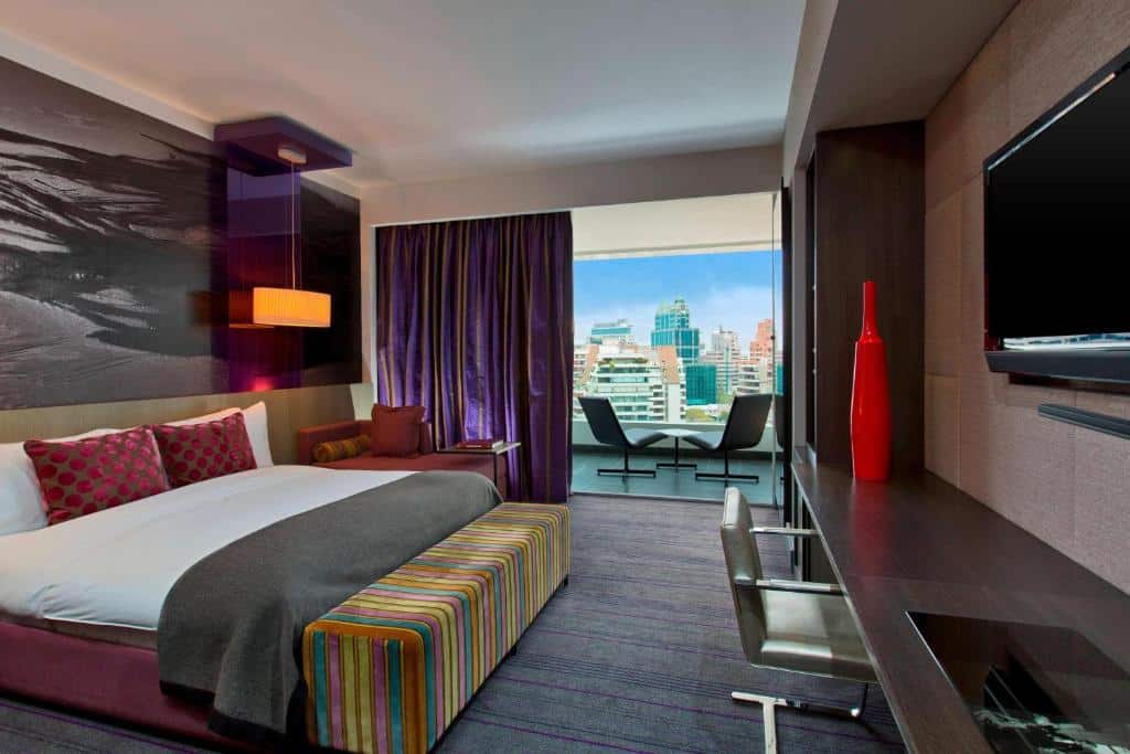 Quarto do W Santiago Hotel com cama de casal do lado esquerdo, cômoda com TV em frente a cama com uma cadeira e do lado esquerdo do quarto um sofá de canto e uma varanda com sacada.