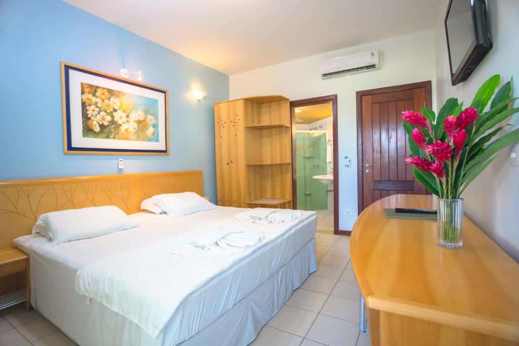 Um quarto do Wetiga Hotel com uma cama de casal, móveis de madeira, uma TV na parede e uma porta de banheiro aberta ao fundo.