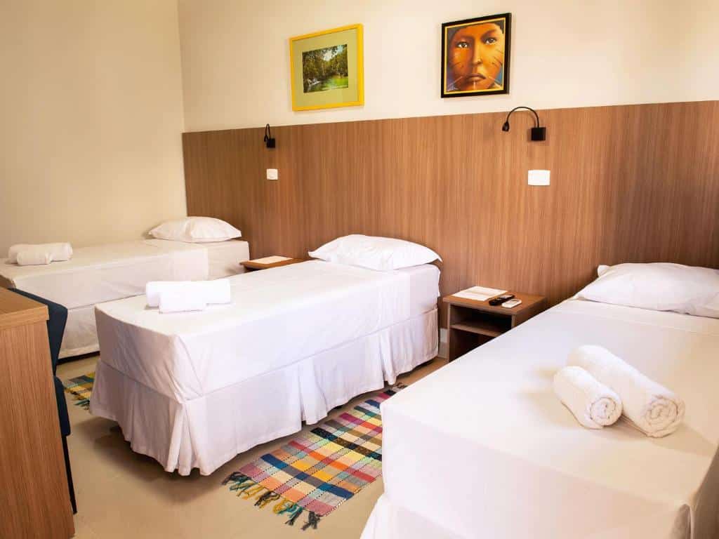 Três camas de solteiro com roupa de cama branca em um quarto do CLH Suites Bonito Centro, dois tapetes coloridos e arte emoldurada em uma parede com painéis de madeira.