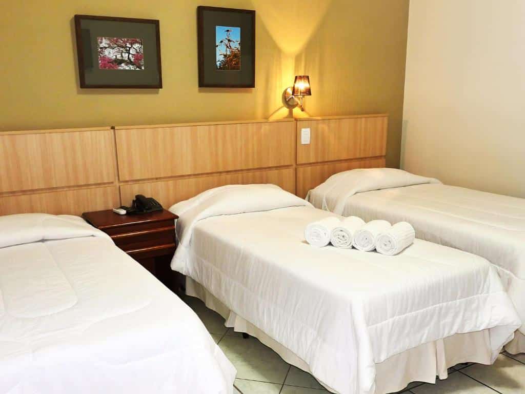 Um quarto do CLH Suites Bonito Sul com três camas de solteiro, roupa de cama branca, toalhas enroladas, uma mesa de cabeceira e dois quadros na parede.