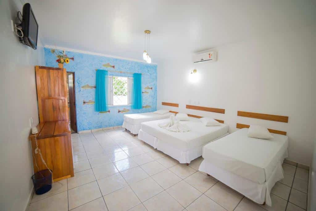 Quarto do Hotel Arauna espaçoso com três camas, parede com detalhes em azul, móveis de madeira, TV e janela com cortinas azuis.