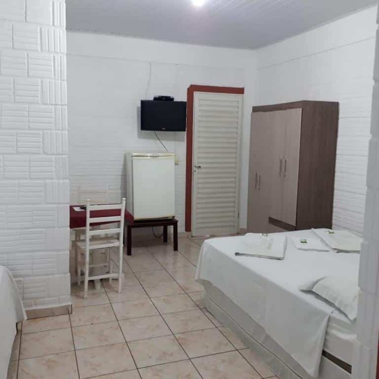 Um quarto simples do Chalé Apart Hotel com cama, guarda-roupa, frigobar pequeno, TV de parede, mesa com cadeiras e piso de azulejo.