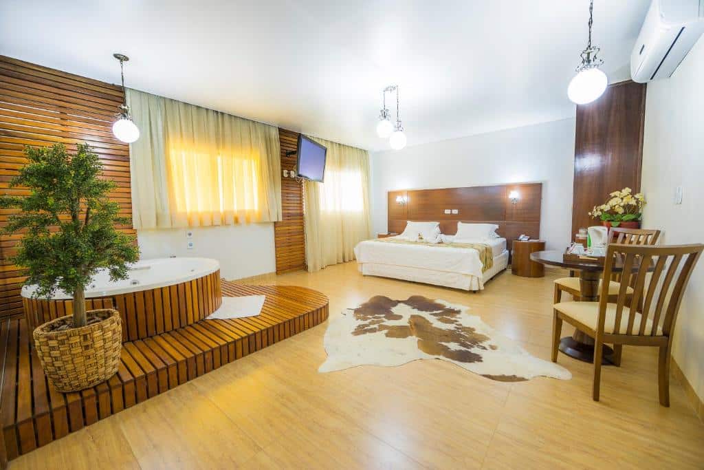Quarto do Marruá Hotel espaçoso com uma cama, banheira de hidromassagem interna, tapete de couro de vaca, detalhes em madeira, uma pequena mesa e um vaso de plantas.