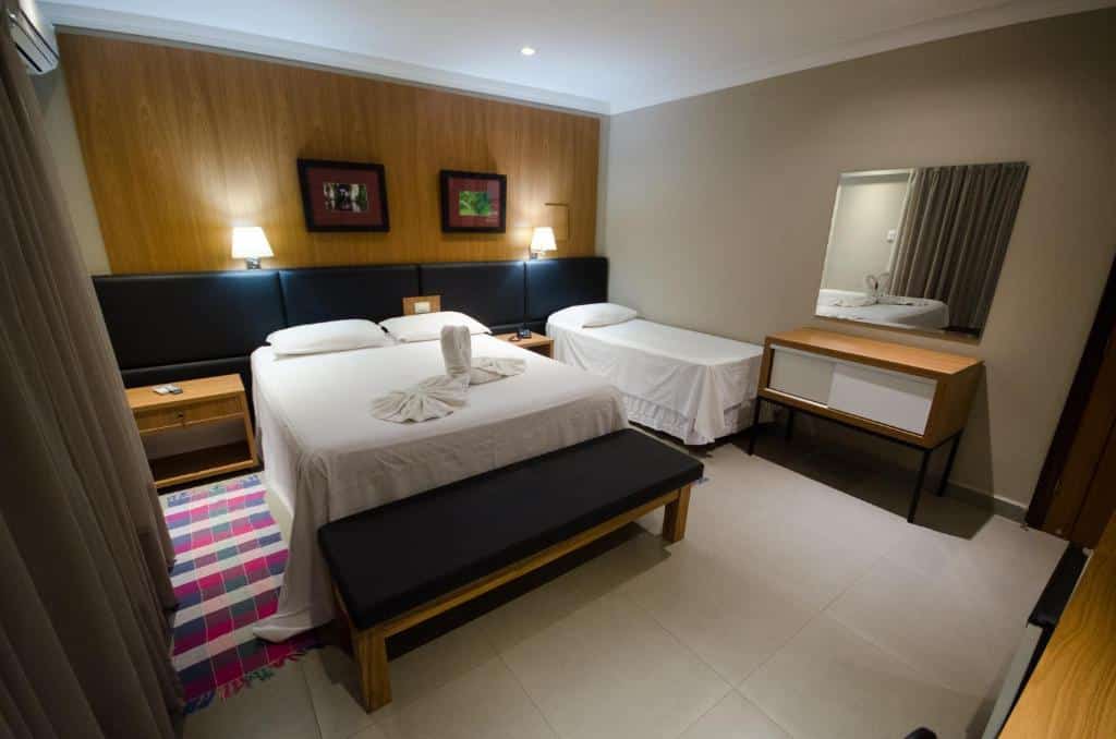 Um quarto do VOA Hotel Paraíso das Águas com uma cama de casal, uma cama de solteiro, uma escrivaninha com espelho e luminárias de parede.
