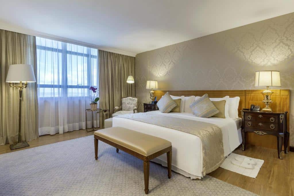 Quarto espaçoso no Meliá Brasil 21 com uma decoração de época, tudo em tons de bege claro e marrom, há uma cama de casal, duas cômodas vintage com abajures, uma janela, uma poltrona e um tapete cinza