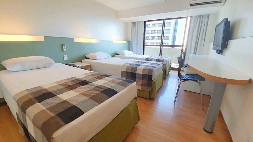 Quarto do St Paul Plaza Hotel com três camas de solteiro, uma sacada, uma televisão, uma mesa de escritório e duas cadeiras, tudo decorado em tons de branco e azul claro, o chão é de madeira
