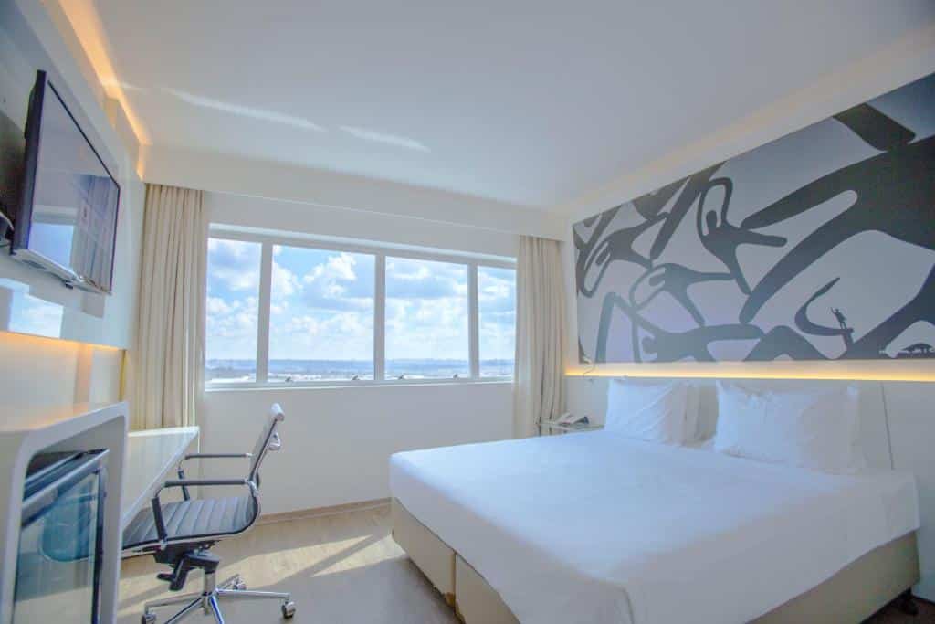 Quarto espaçoso no ibis Styles Brasilia Aeroporto com uma janela dando vista para o lago, uma cama de casal, um frigobar, uma televisão, uma mesa de escritório com cadeira, tudo em tons de branco, para representar hotéis em Brasília