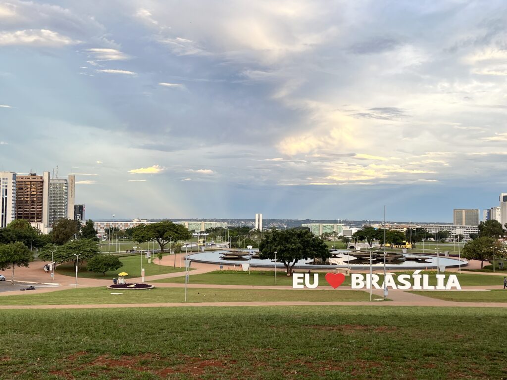 Vista de um parque em Brasília com edifícios ao fundo e uma placa com a inscrição "EU ❤️ BRASÍLIA" em primeiro plano.