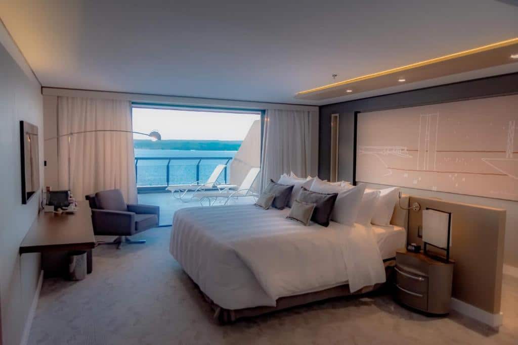Quarto no Royal Tulip Brasília Alvorada com uma sacada ampla com vista para o lago, uma cama de casal, uma poltrona, uma televisão, tudo em tons de bege e marrom claro, para representar hotéis em Brasília