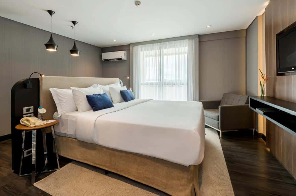Quarto no Grand Mercure Brasilia Eixo Monumental com uma sacada, uma cama de casal, uma poltrona, uma televisão, um ar-condicionado, tudo em tons de madeira, cinza e bege, para representar hotéis em Brasília