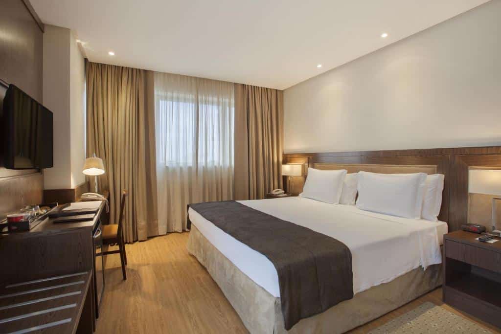 Quarto no Windsor Brasilia Hotel com uma janela, uma cama de casal, uma cabeceira com abajures, uma televisão, uma mesa de escritório com uma cadeira, o chão é de madeira, tudo é decorado num tom de madeira claro
