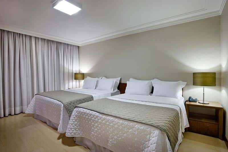 Quarto do Sia Park Executive Hotel com duas camas de casal, duas mesinhas de cabeceira com abajures sob elas, as camas estão com roupas brancas e cinzas, o chão é de madeira, para representar hotéis em Brasília
