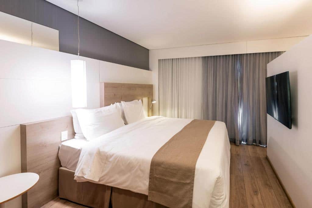 Quarto no Manhattan Plaza com chão de madeira, uma televisão, uma cama de casal, tudo em tons de marrom e branco