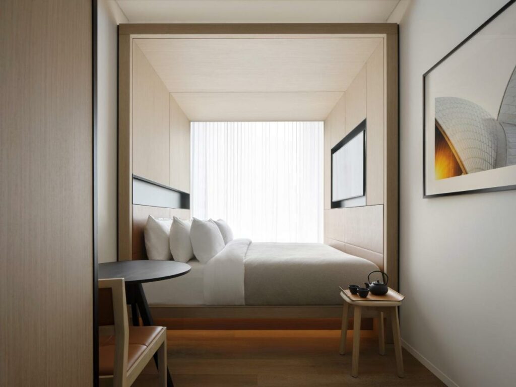 Quarto do Little National Hotel. Do lado esquerdo uma mesa com uma poltrona, do lado direito um banco, no fundo a cama com TV e atrás uma janela. Imagem para ilustrar post sobre hotéis em Sydney.