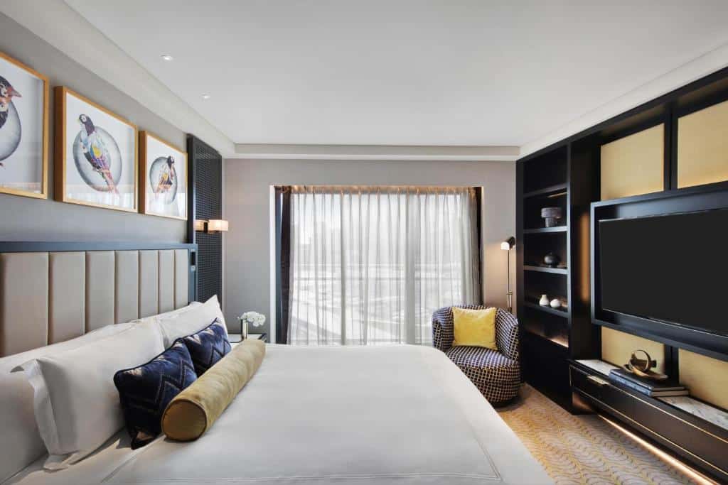 Quarto do The Star Grand Hotel and Residences. Do lado esquerdo uma mesa de casal, no fundo uma prateleira e um lustre. Do lado direito um painel com prateleiras, uma poltrona e um abajur. No fundo uma parede de vidro. 