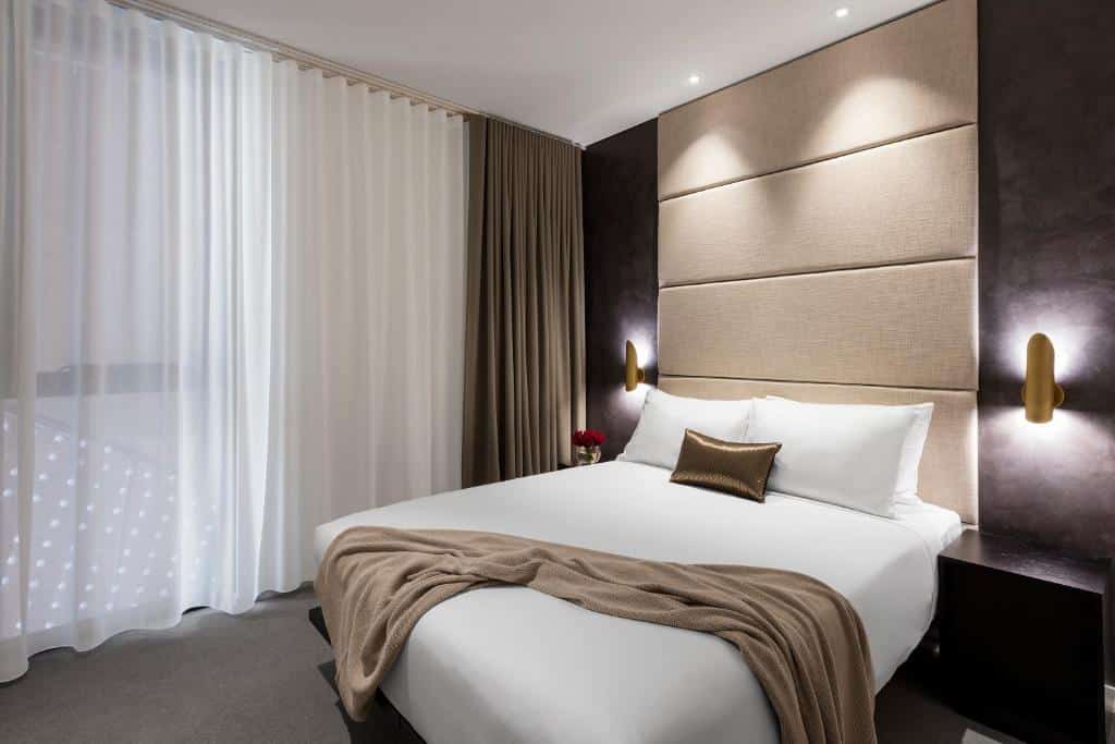 Quarto do SKYE Suites. Uma cama de casal do lado direito, com uma cômoda e um lustre de cada lado. Do lado esquerdo uma parede de vidro com janelas. 